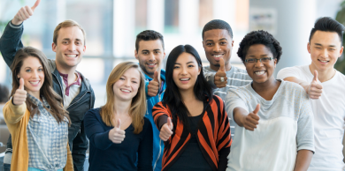 eight students of a variety of ethnicities and genders posing outside holding their thumbs up